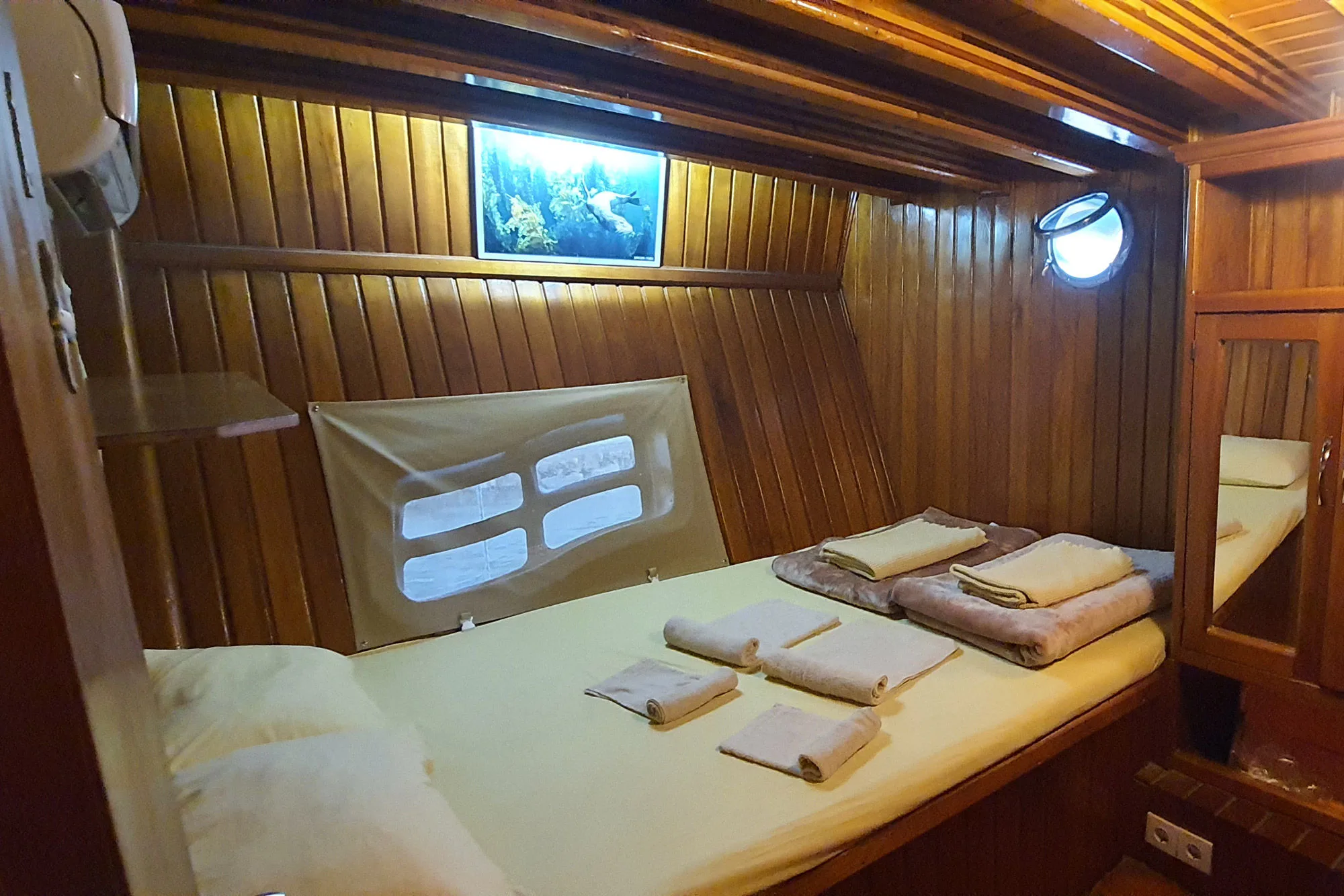 Timber-lined Okyanus JD cabin with a double bed, two portholes and a shelf above the headboard