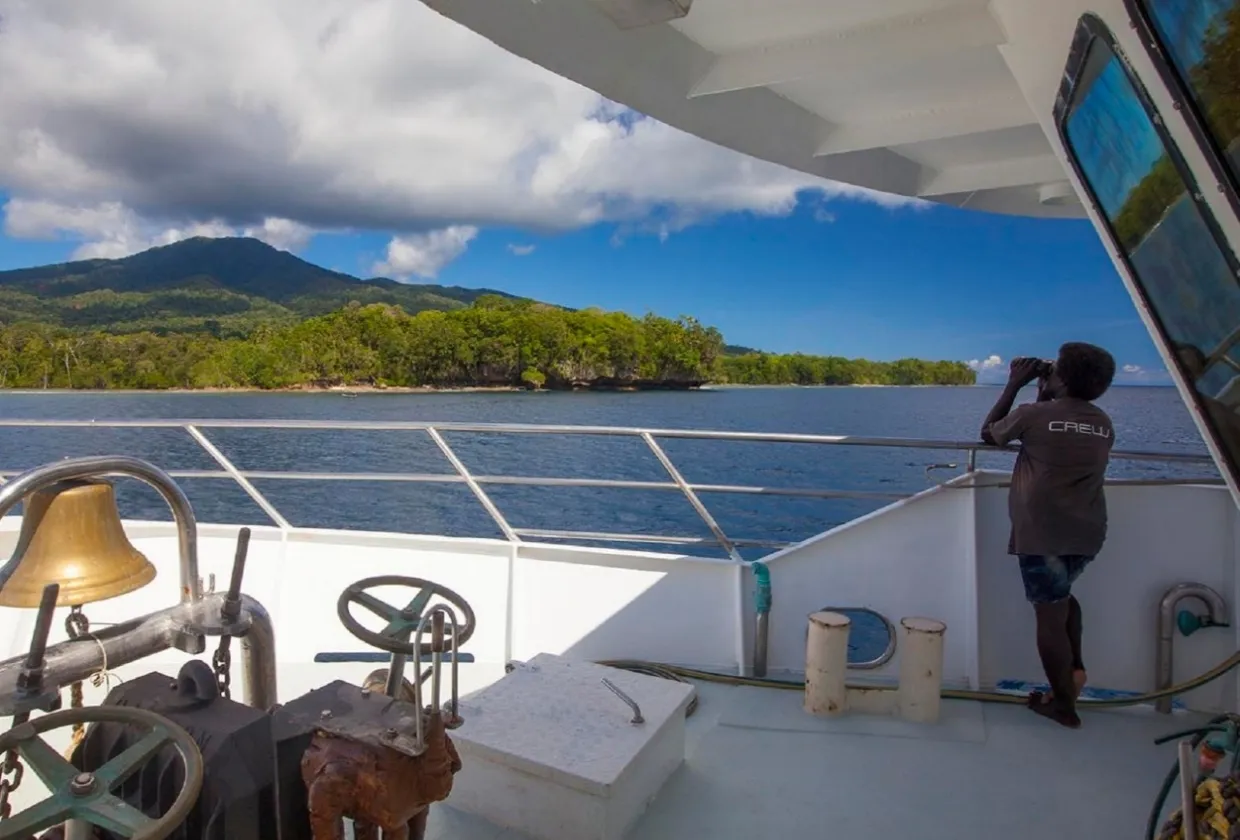 Pacific Master anchored off a green island, crew member on watch at the bow