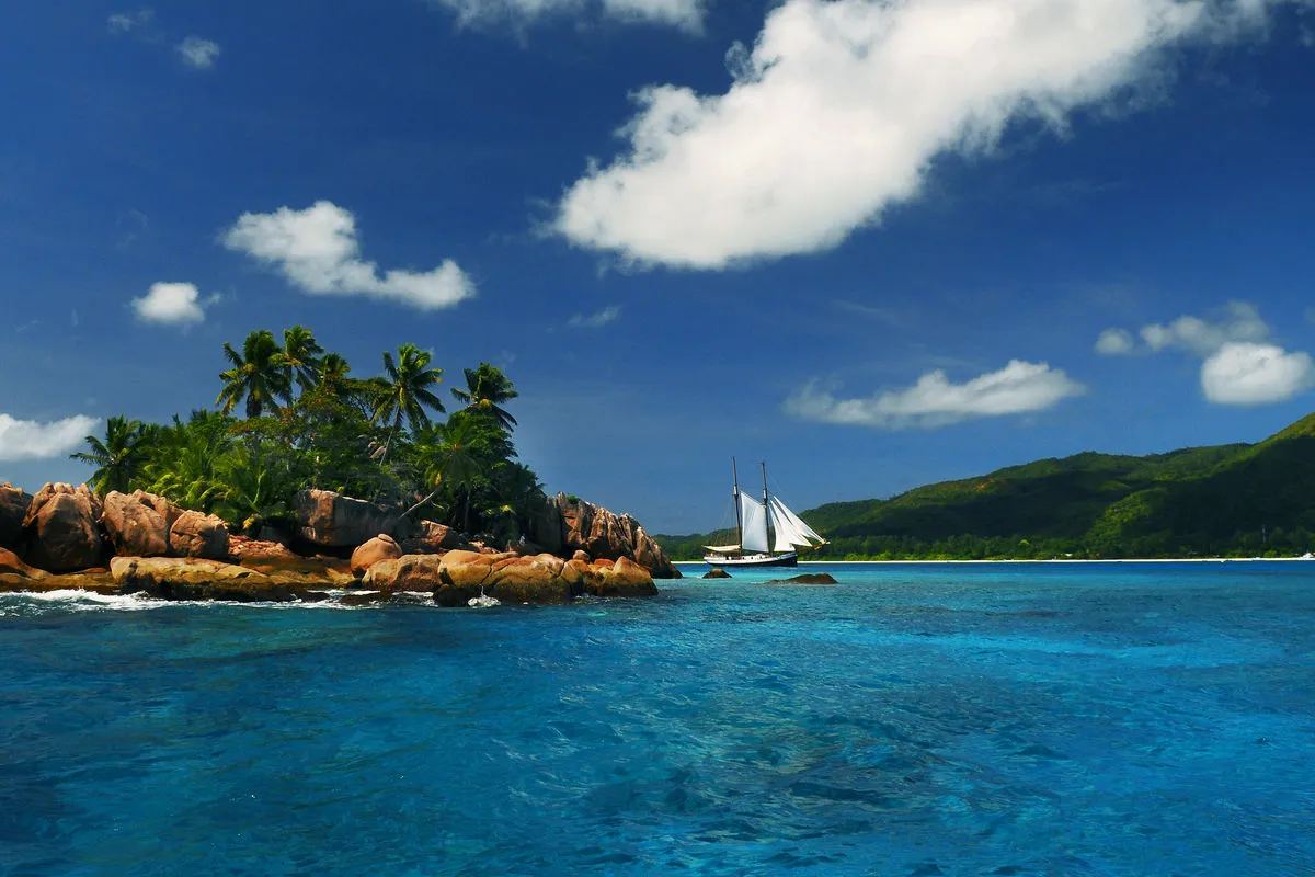 A white sail in the distance over a turquoise lagoon strewn with granite boulders